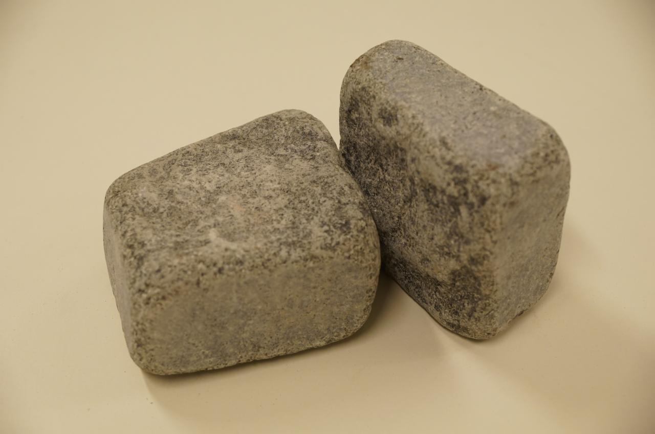 Dwie szare kostki granitowe o nieregularnych kształtach na beżowym tle. Widoczna faktura kamienia i delikatne cieniowanie. Ujęcie z bliska, ukazujące detale.