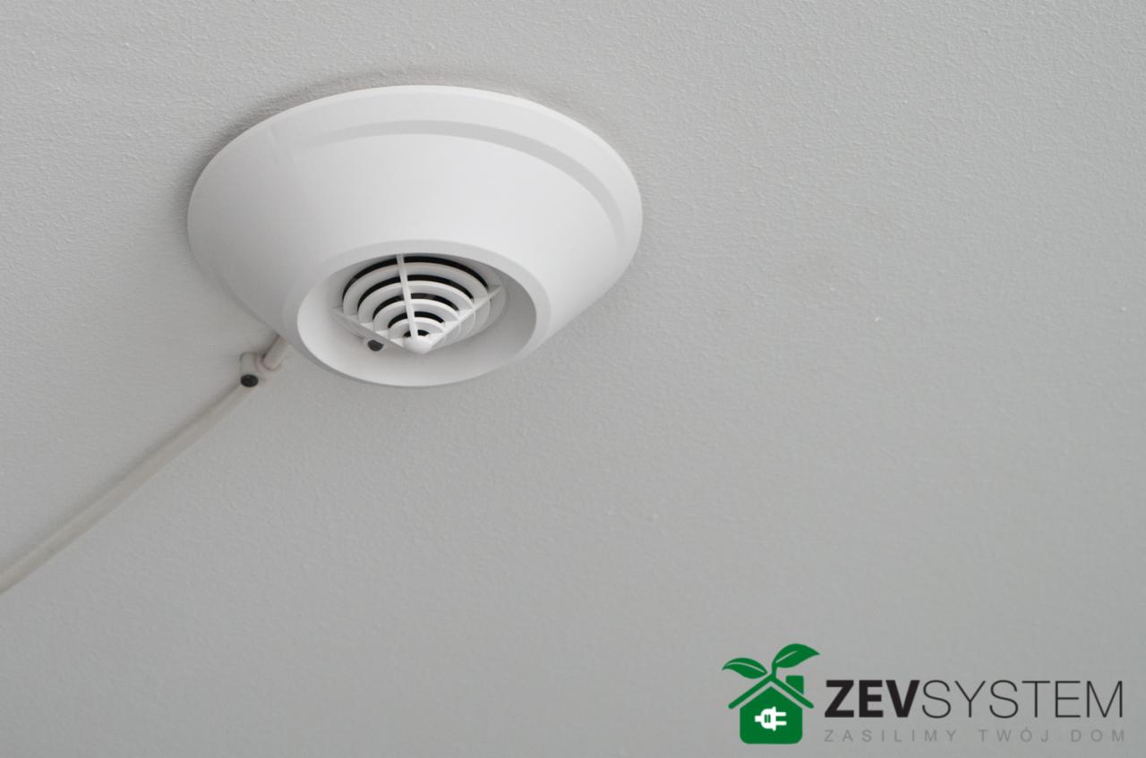 Biały czujnik systemu alarmowego, prawdopodobnie dymu, zamontowany na białym suficie, z widocznym kablem i uchwytem kabla. W prawym dolnym rogu logo firmy Zevsystem.