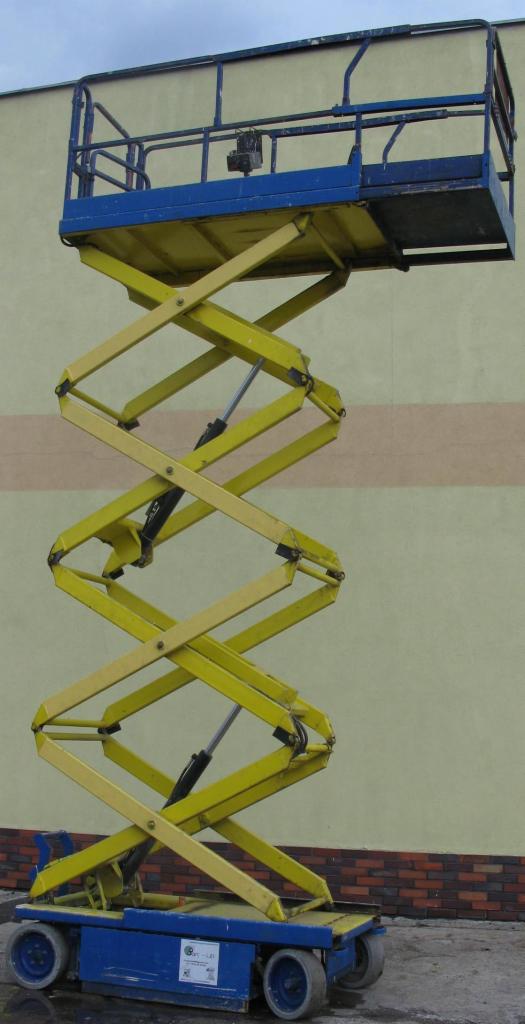 Żółto-niebieski podnośnik nożycowy na tle ściany z kremowym i brązowym pasem, widok z boku, platforma robocza z barierkami.
