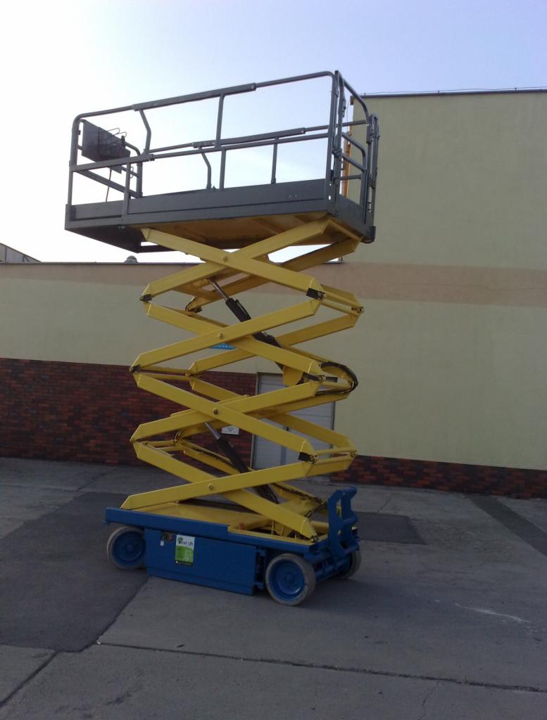 Żółto-niebieski podnośnik nożycowy na betonowej powierzchni przed budynkiem z beżową i ceglaną elewacją w słoneczny dzień.