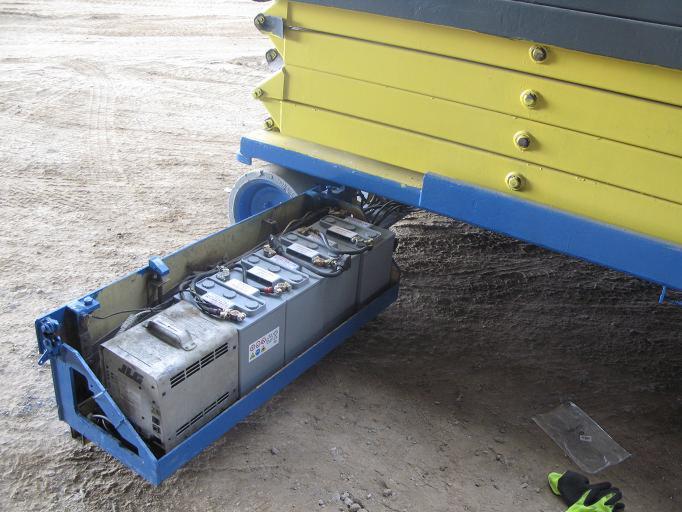 Szczegółowy widok akumulatorów trakcyjnych w niebieskiej metalowej obudowie pod żółtym podnośnikiem nożycowym JLG na szarym, betonowym podłożu. Widoczne przewody, naklejki ostrzegawcze i fragment...