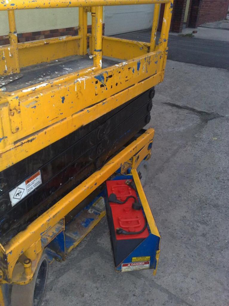 Żółty podnośnik koszowy z widocznym akumulatorem trakcyjnym (bateria trakcyjna) w czerwonej obudowie, stojący na betonowej powierzchni.