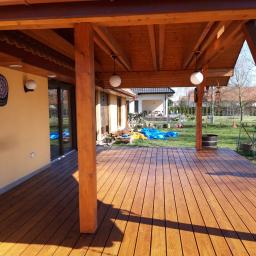 Dawid Krupka - Zadaszony taras z drewnianą podłogą i słupami, widoczne oświetlenie w postaci kulistych lamp, dach pokryty czerwoną dachówką, w tle dom i ogród.