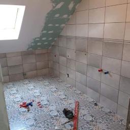 Dawid Krupka - Łazienka w trakcie remontu na poddaszu: ułożone płytki na podłodze z wzorem, częściowo wykończone ściany z szarymi płytkami, fragment sufitu skośnego z nałożoną gładzią, widoczne rury instalacji...