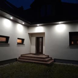 Dawid Krupka - Elewacja domu z oświetleniem zewnętrznym, schody wejściowe z brązowych płytek prowadzące do ciemnych drzwi wejściowych, małe okna z ciemnymi ramami, widok nocny.