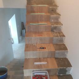 Dawid Krupka - Montaż płytek imitujących drewno na schodach w Odolanowie, widoczne poziomice, krzyżyki dystansowe i pojemnik z klinami poziomującymi.