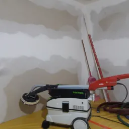 Szlifowanie ściany pokrytej gładzią za pomocą szlifierki Festool Planex LHS 225 z odkurzaczem, widoczne nierówności na ścianie i narzędzia w tle.