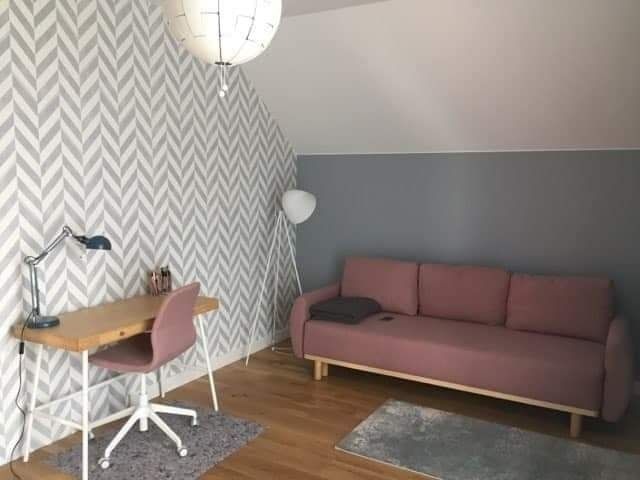 Pokój z biurkiem, lampką, krzesłem i różową sofą na tle ściany w zygzaki i szarej ściany pod skosem dachu.