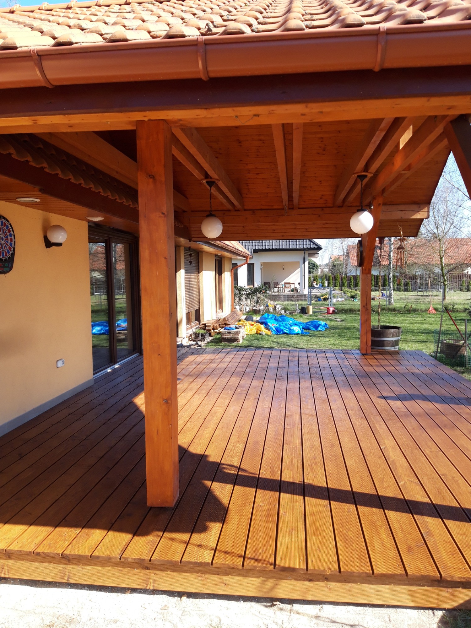 Zadaszony taras z drewnianą podłogą i słupami, widoczne oświetlenie w postaci kulistych lamp, dach pokryty czerwoną dachówką, w tle dom i ogród.