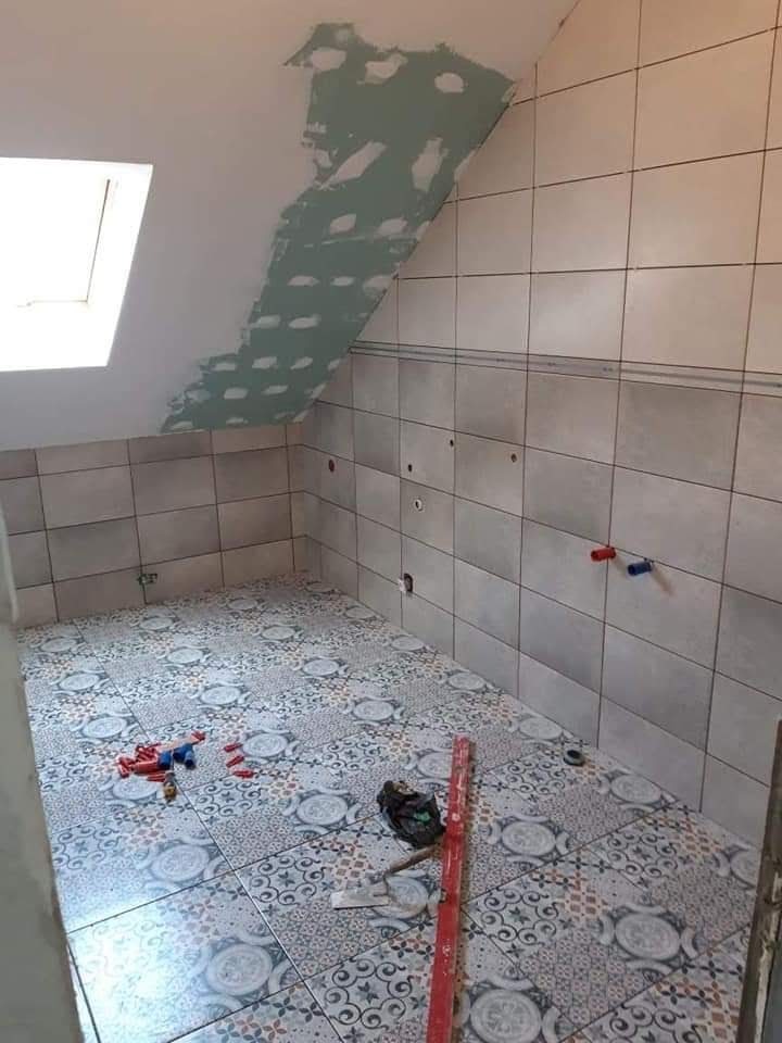 Łazienka w trakcie remontu na poddaszu: ułożone płytki na podłodze z wzorem, częściowo wykończone ściany z szarymi płytkami, fragment sufitu skośnego z nałożoną gładzią, widoczne rury instalacji...