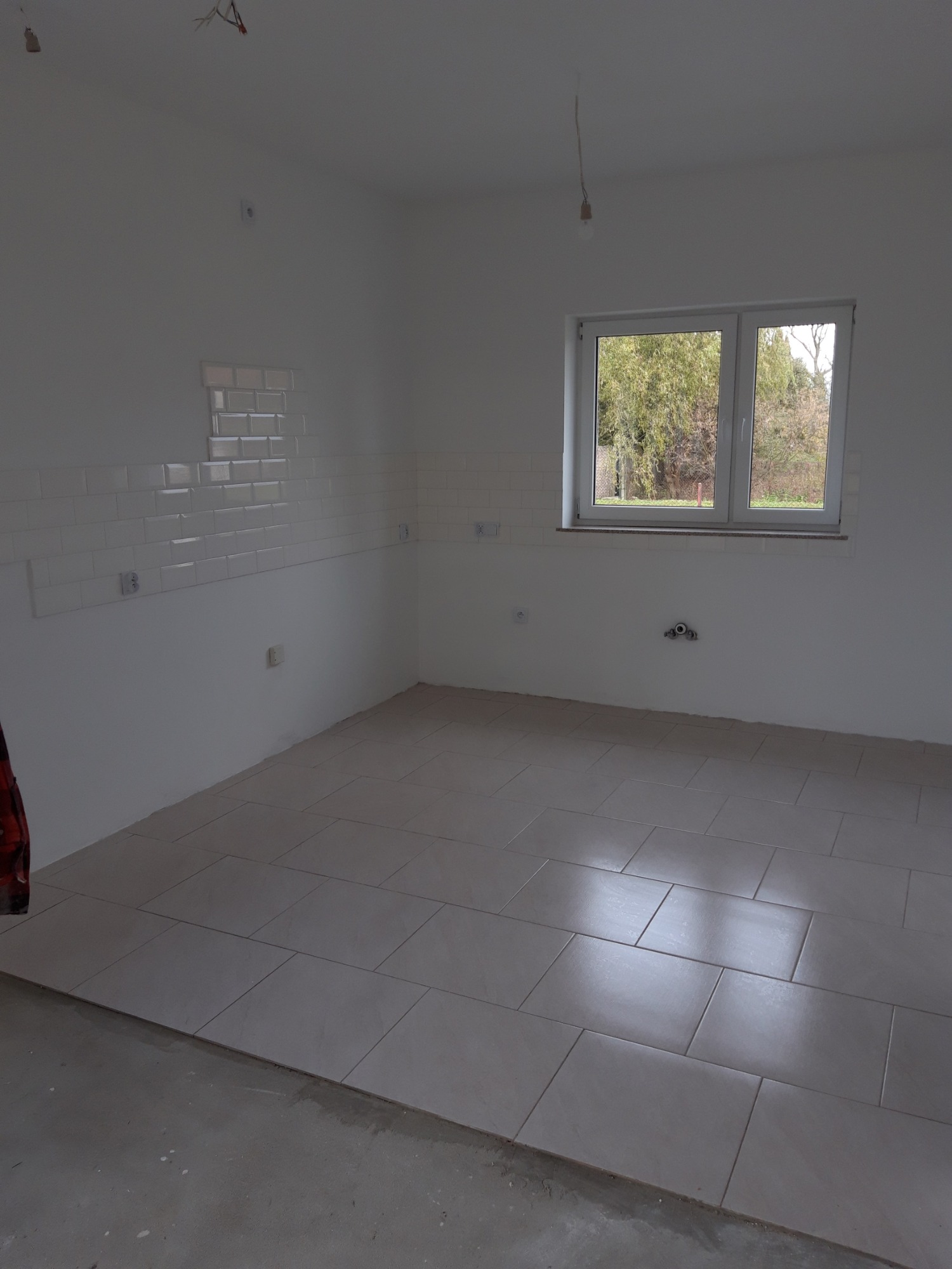 Wnętrze w trakcie prac wykończeniowych: częściowo ułożone płytki podłogowe w odcieniach szarości, fragment ściany z białymi, błyszczącymi płytkami typu metro, okno z widokiem na drzewa, surowy stan...