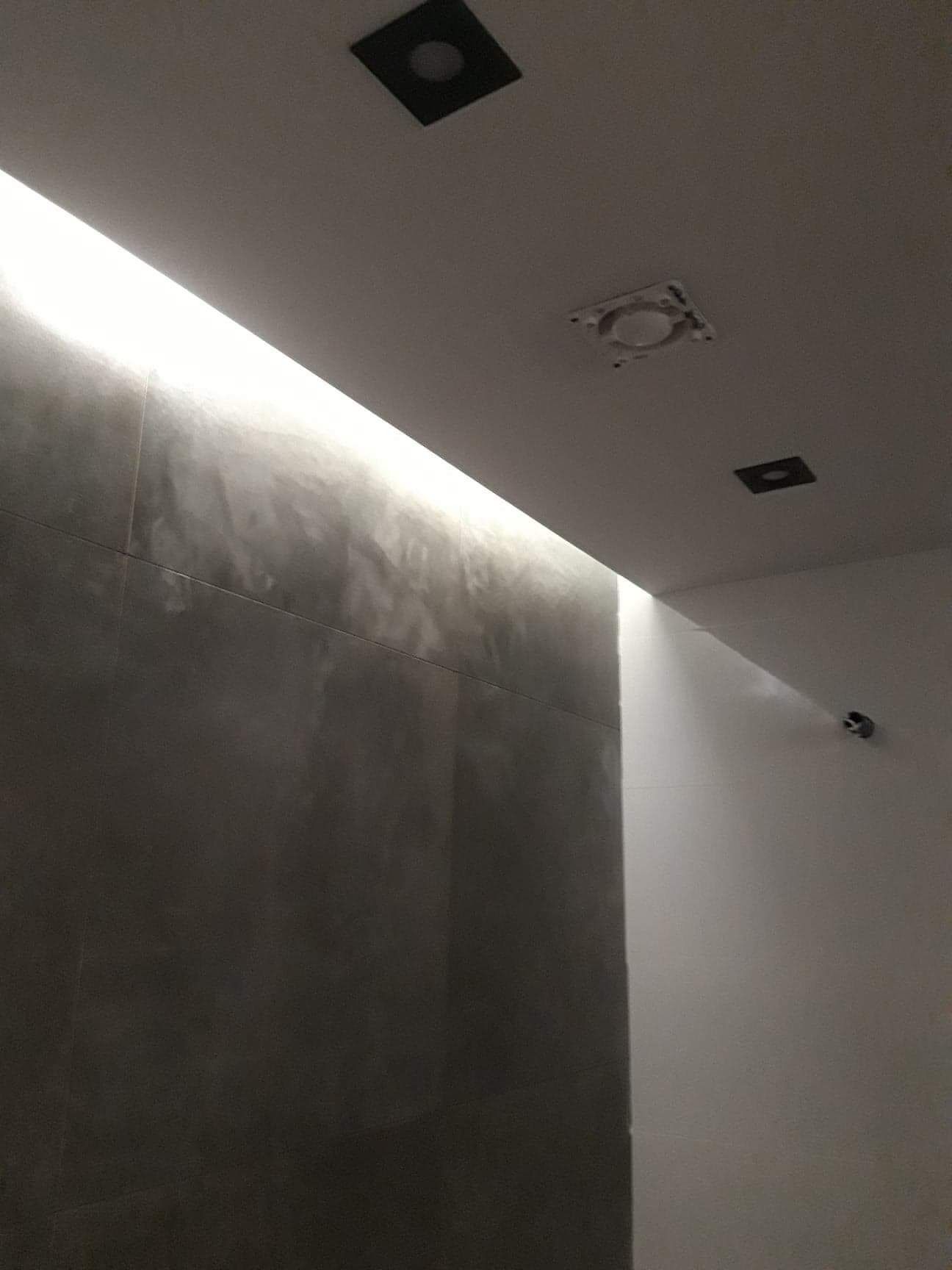 Ściana łazienki wyłożona szarymi płytkami z efektem betonu, oświetlona listwą LED w suficie, widoczne także czarne oprawy oświetleniowe i instalacja pod punkt świetlny.