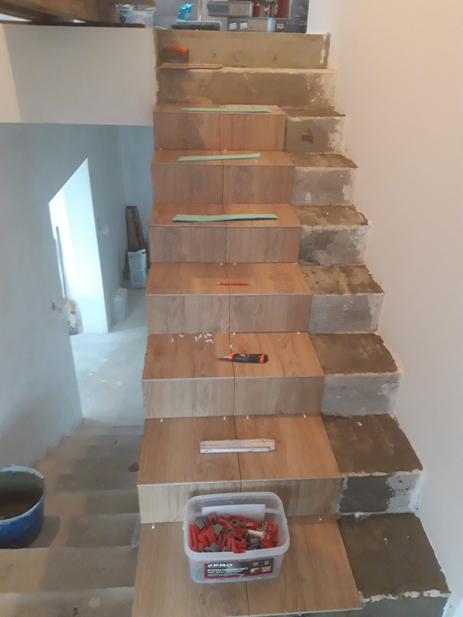 Montaż płytek imitujących drewno na schodach w Odolanowie, widoczne poziomice, krzyżyki dystansowe i pojemnik z klinami poziomującymi.