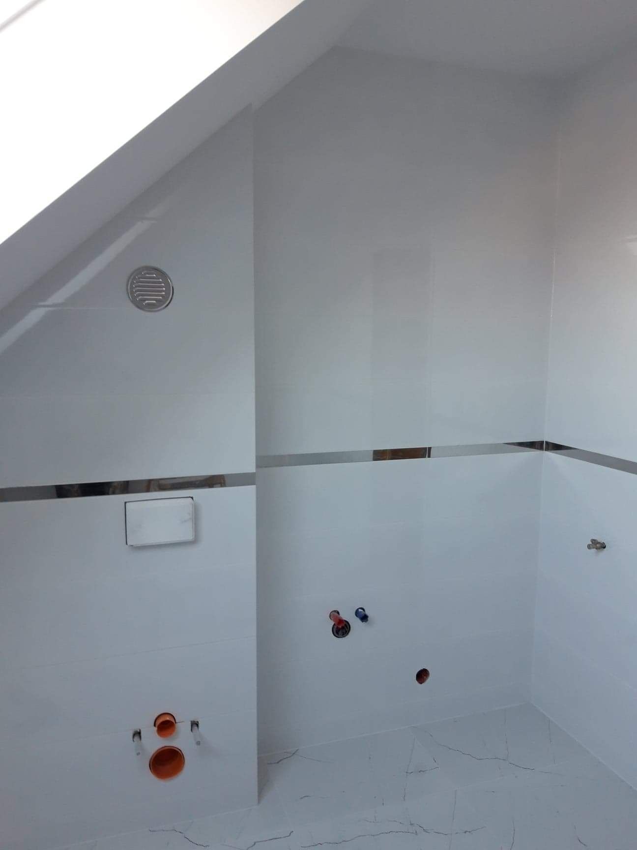 Biała łazienka w trakcie remontu, widoczne białe płytki z metalicznymi paskami, otwory na armaturę i odpływ, wentylacja pod skosem dachu, podłoga z marmurkowym wzorem.