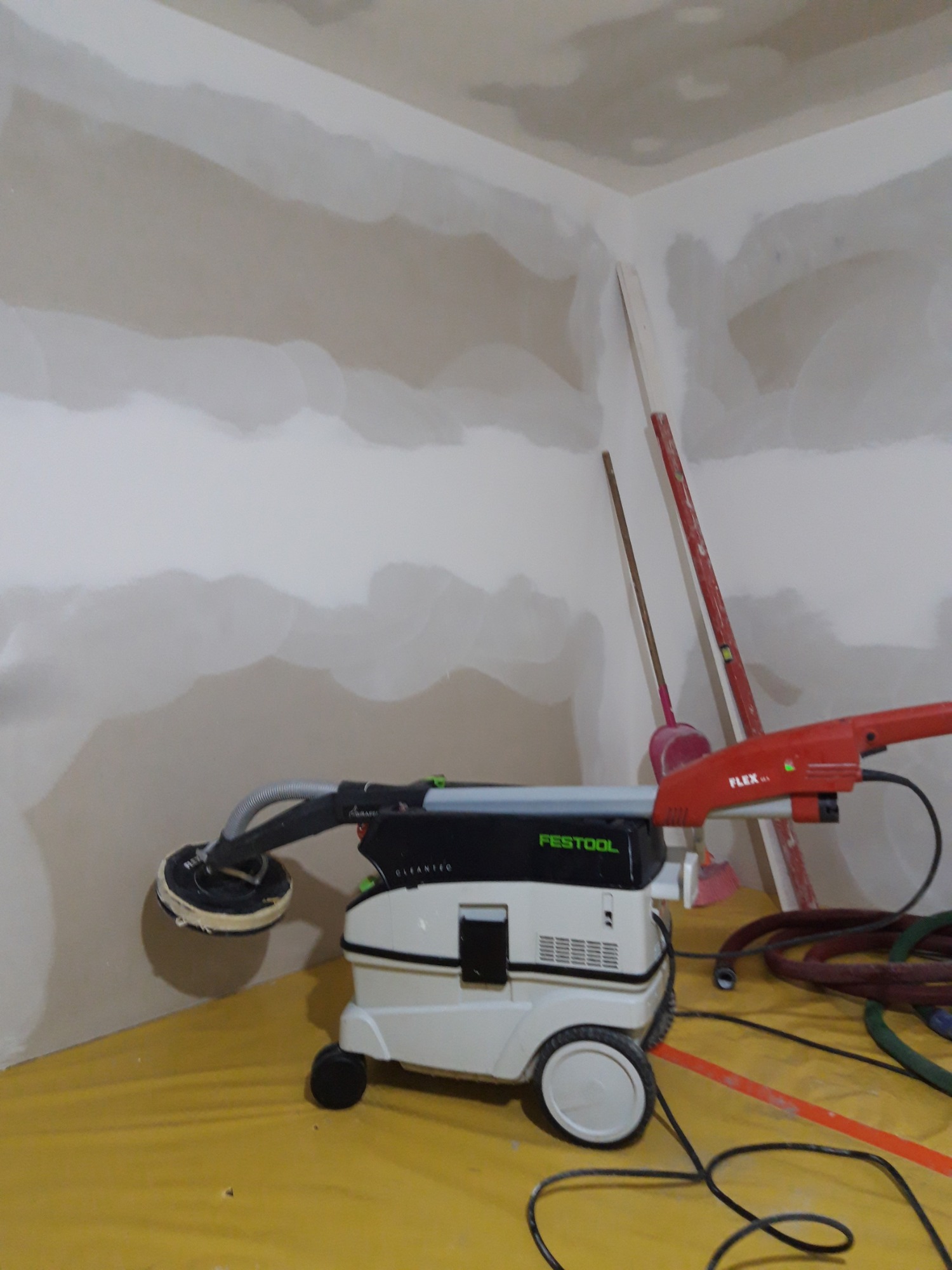 Szlifowanie ściany pokrytej gładzią za pomocą szlifierki Festool Planex LHS 225 z odkurzaczem, widoczne nierówności na ścianie i narzędzia w tle.