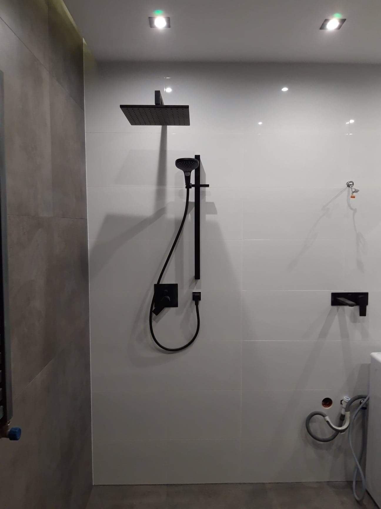 Nowoczesny zestaw prysznicowy w kolorze czarnym, zamontowany na białych, błyszczących płytkach w łazience. Widoczne przyłącza wodne i odpływowe w ścianie, sugerujące etap prac wykończeniowych.