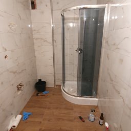 Krzysztof Kondrat - Montaż kabiny prysznicowej narożnej w łazience z płytkami imitującymi marmur, widoczne narzędzia i chemia budowlana na podłodze z paneli winylowych.