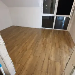 Ułożona podłoga z płytek imitujących drewno w jasnym pomieszczeniu z dużym oknem, widok przez surowy otwór drzwiowy z widocznymi śladami zaprawy.