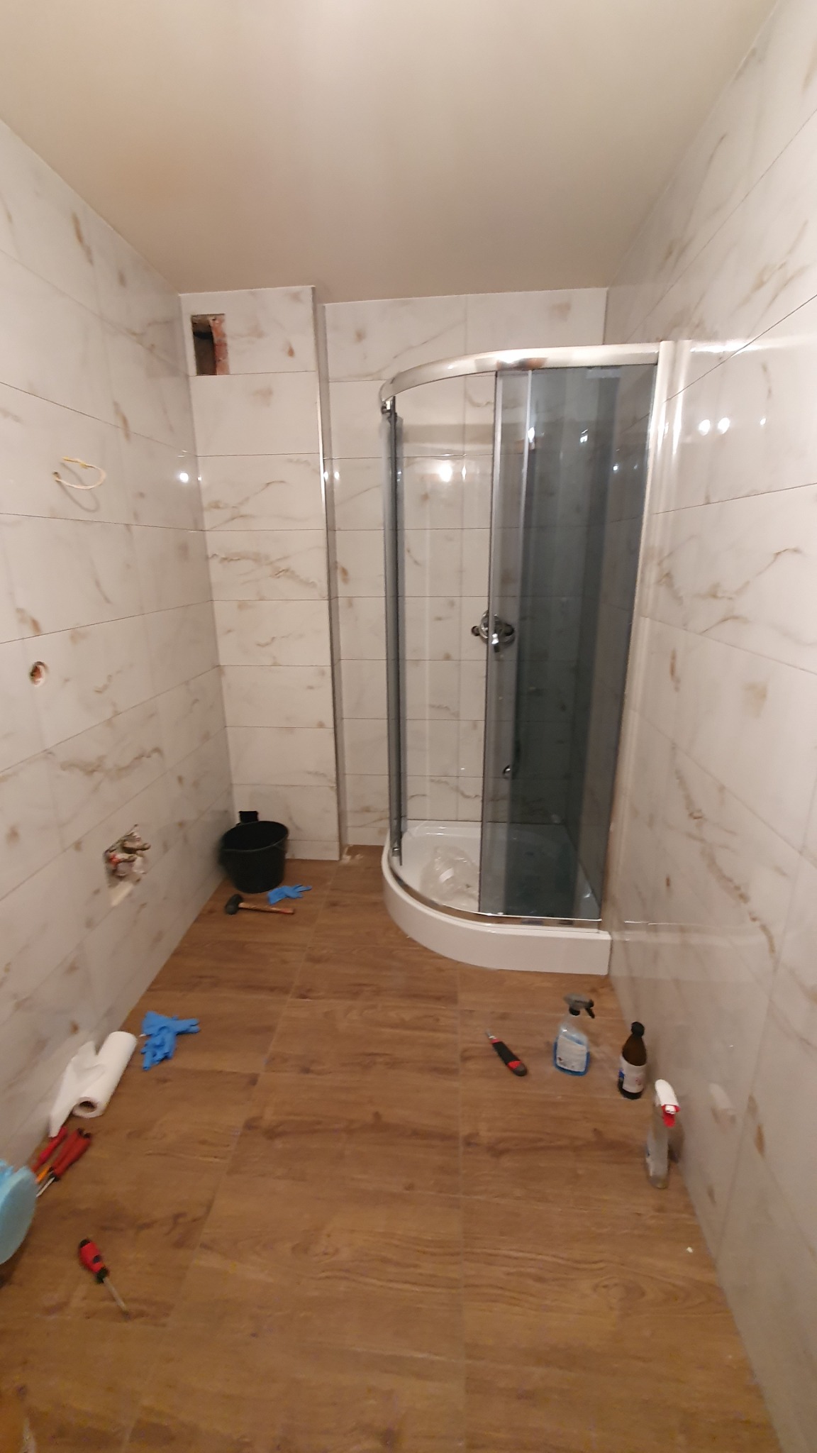 Montaż kabiny prysznicowej narożnej w łazience z płytkami imitującymi marmur, widoczne narzędzia i chemia budowlana na podłodze z paneli winylowych.