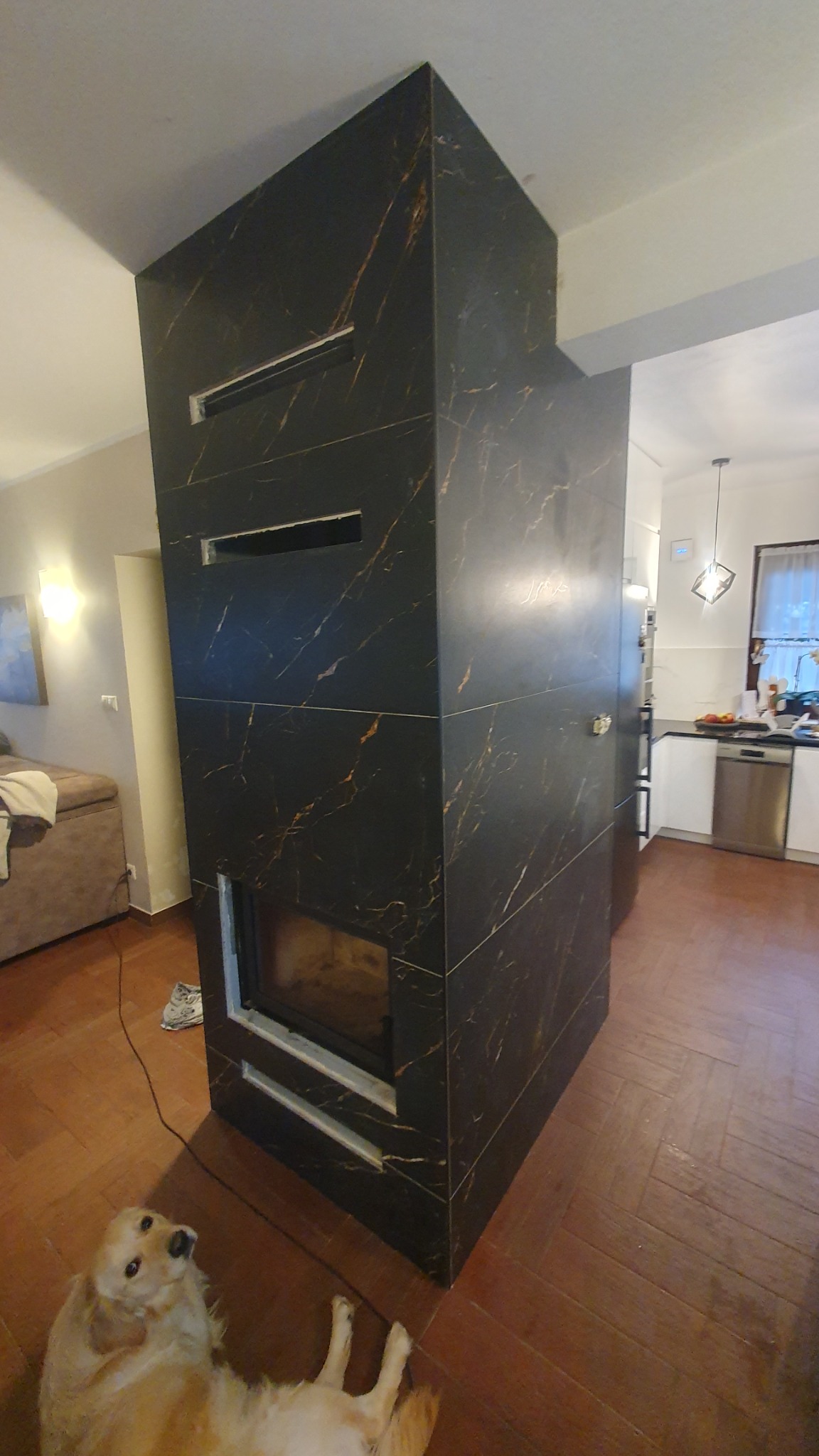 Obudowa kominka wykończona czarnymi płytkami z marmurkowym wzorem, widoczny fragment salonu i kuchni, na pierwszym planie leżący pies rasy golden retriever.