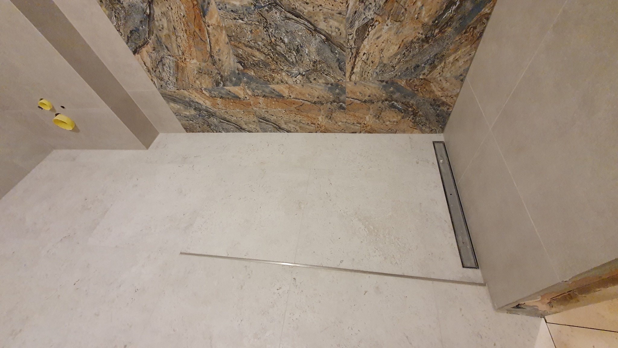 Wykończona łazienka z beżowymi płytkami podłogowymi i ściennymi, z dekoracyjnym panelem imitującym kamień na suficie nad prysznicem, widoczny odpływ liniowy