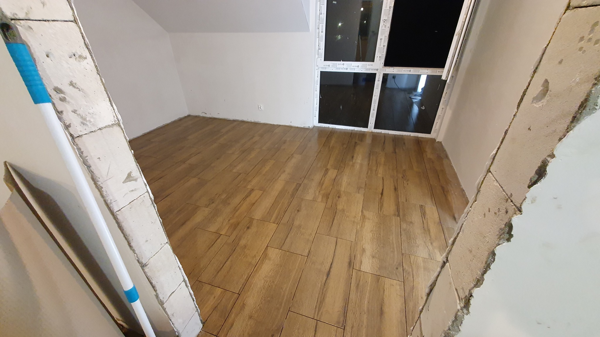 Ułożona podłoga z płytek imitujących drewno w jasnym pomieszczeniu z dużym oknem, widok przez surowy otwór drzwiowy z widocznymi śladami zaprawy.