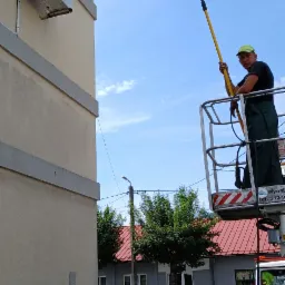 Pracownik w zielonym kombinezonie i czarnej koszulce, stojący na podnośniku koszowym, trzyma lancę do czyszczenia elewacji budynku z klimatyzatorami, widok na błękitne niebo i budynki w tle.