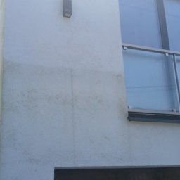 Clean Service - Boczna ściana budynku z widocznymi śladami zabrudzeń i zacieków na elewacji, fragment okna z barierką i brama garażowa z ciemnego drewna.