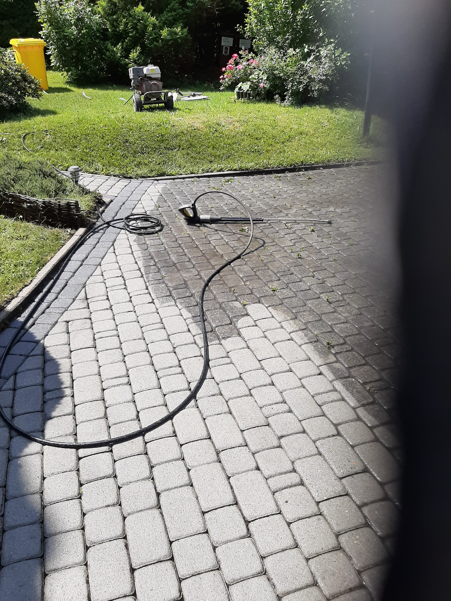 Częściowo umyta kostka brukowa na podjeździe, widoczny wąż ciśnieniowy i urządzenie czyszczące na trawniku w tle.