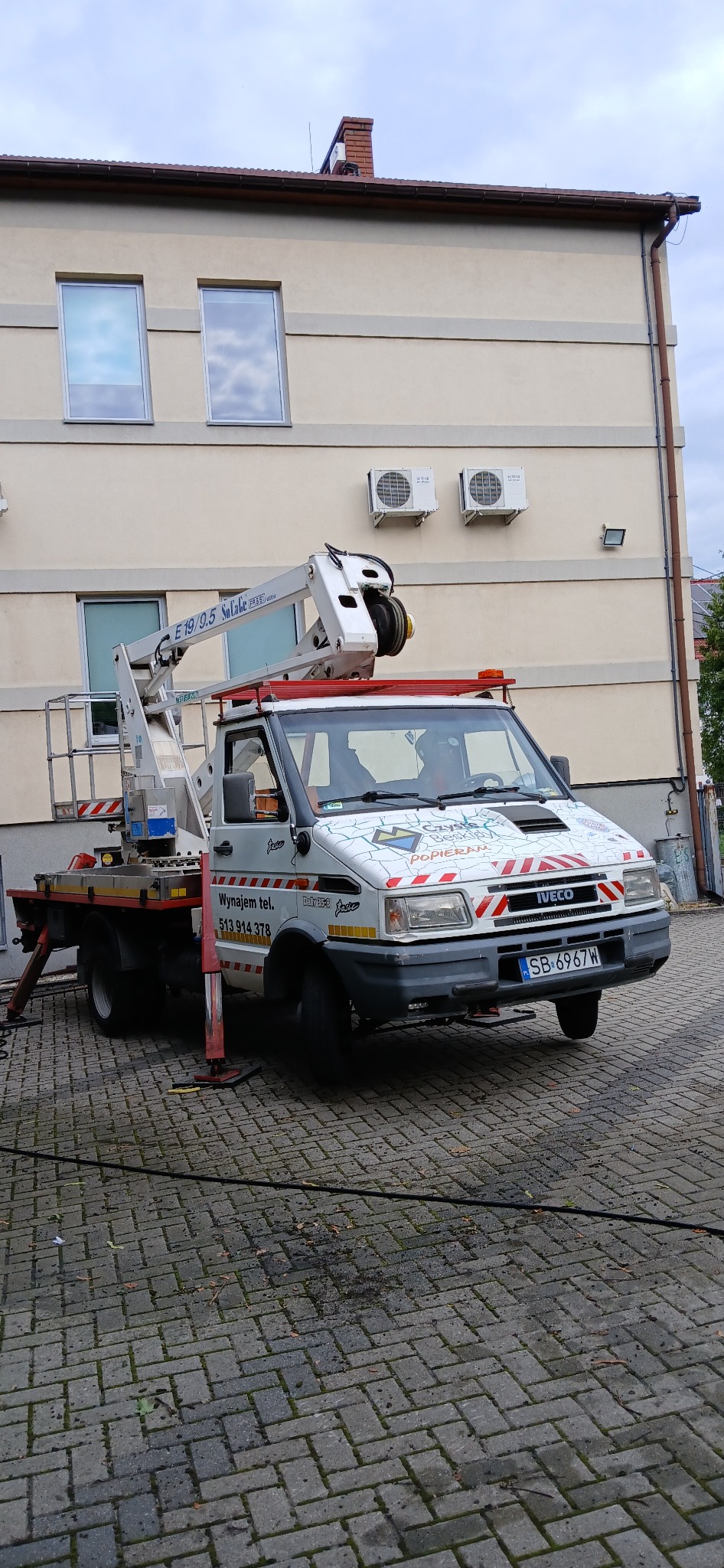 Podnośnik koszowy Iveco SoLate E19/9.5 zaparkowany na kostce brukowej przed budynkiem z klimatyzacją, podpory stabilizujące rozłożone.
