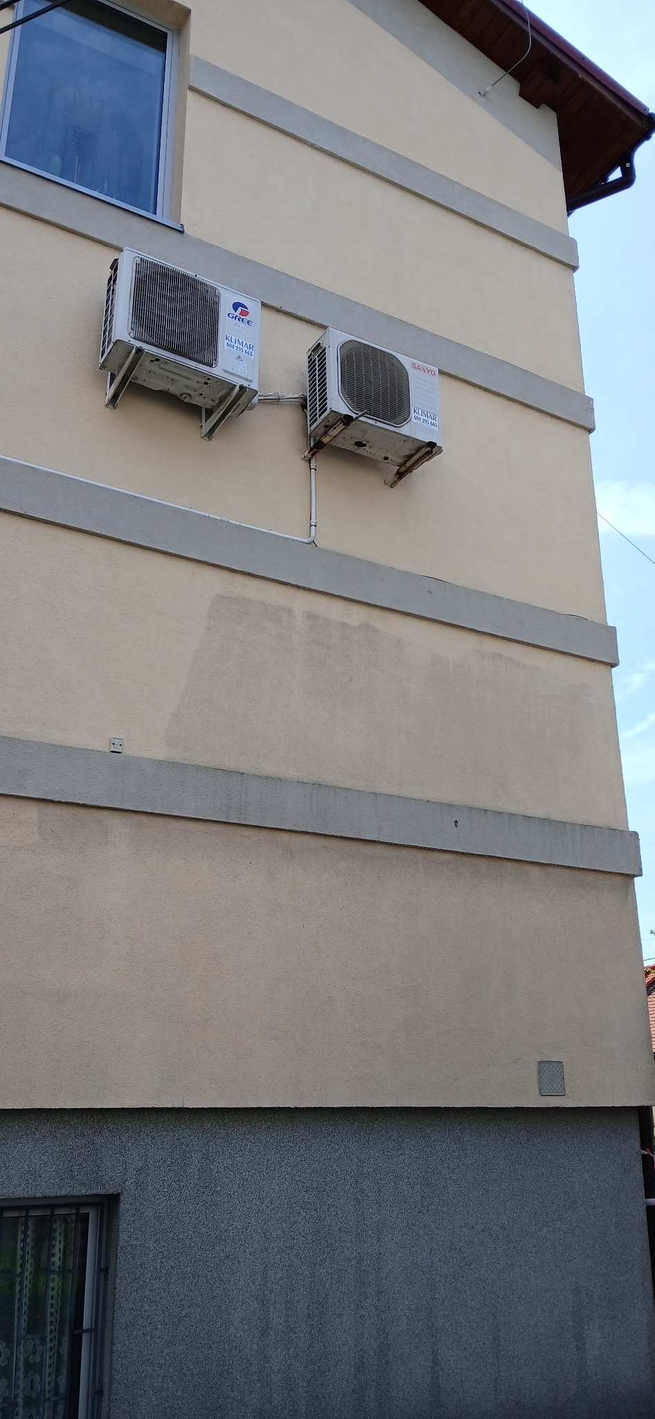 Dwa klimatyzatory różnych marek (Gree i Sanyo) zamontowane na beżowej elewacji budynku z szarymi pasami dekoracyjnymi i widocznymi śladami po czyszczeniu elewacji.