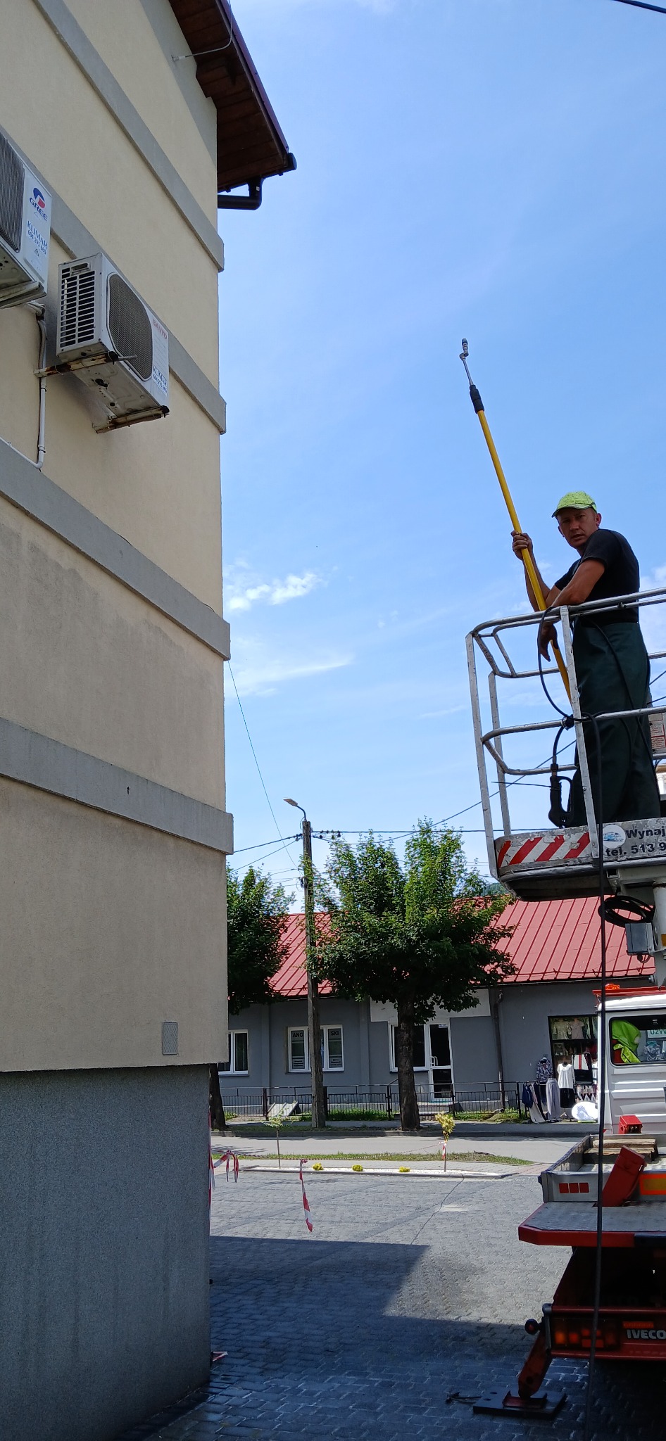 Pracownik w zielonym kombinezonie i czarnej koszulce, stojący na podnośniku koszowym, trzyma lancę do czyszczenia elewacji budynku z klimatyzatorami, widok na błękitne niebo i budynki w tle.