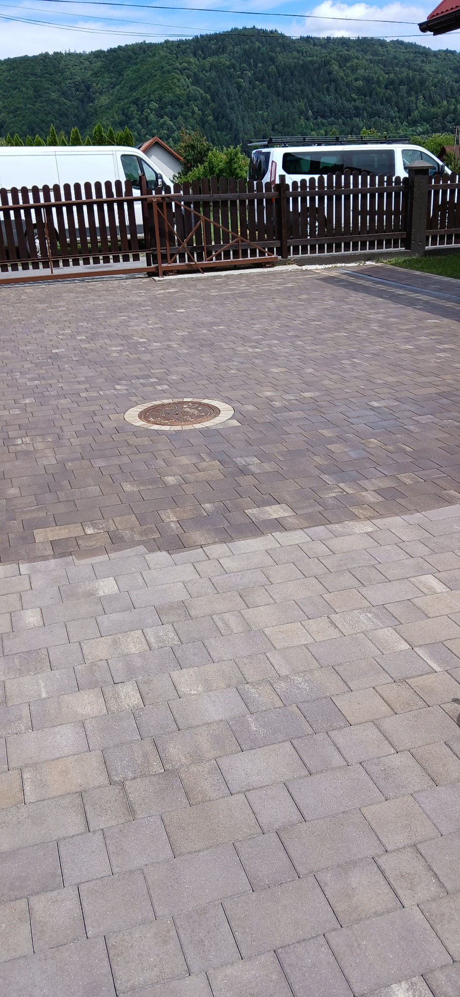 Fragment kostki brukowej, część wyczyszczona, widoczna różnica w kolorze, studzienka kanalizacyjna, ogrodzenie z bramą, dwa samochody dostawcze w tle, zalesione wzgórze.