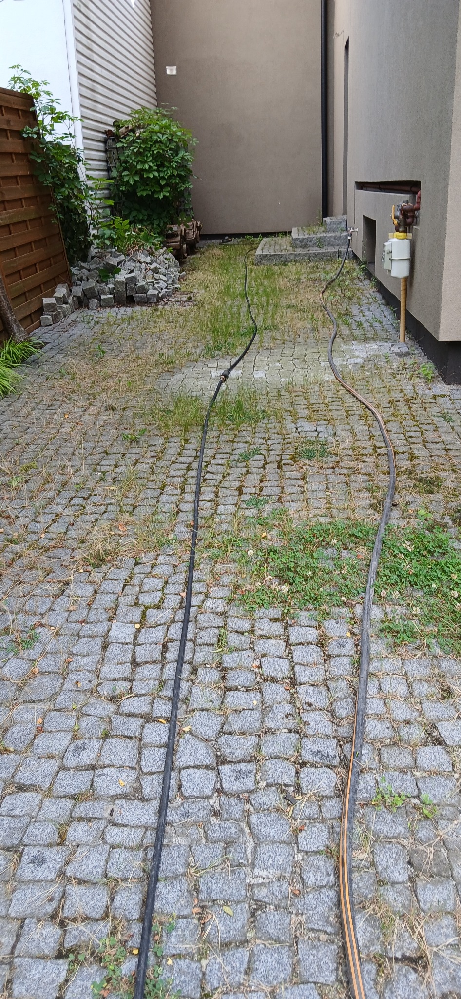 Dwie węże ciśnieniowe leżące na zaniedbanej kostce brukowej z chwastami, prowadzące w kierunku ściany budynku i schodów.