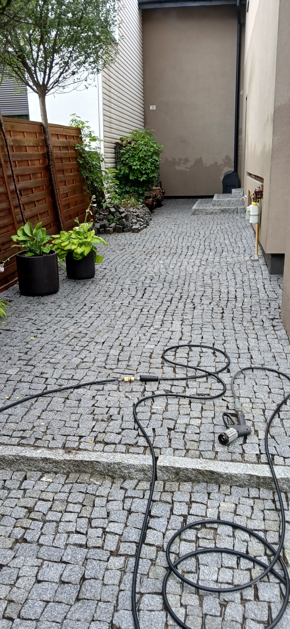 Szara kostka brukowa na posesji z widoczną lancą do mycia pod ciśnieniem i wężem, donice z roślinami i drewniane ogrodzenie w tle.