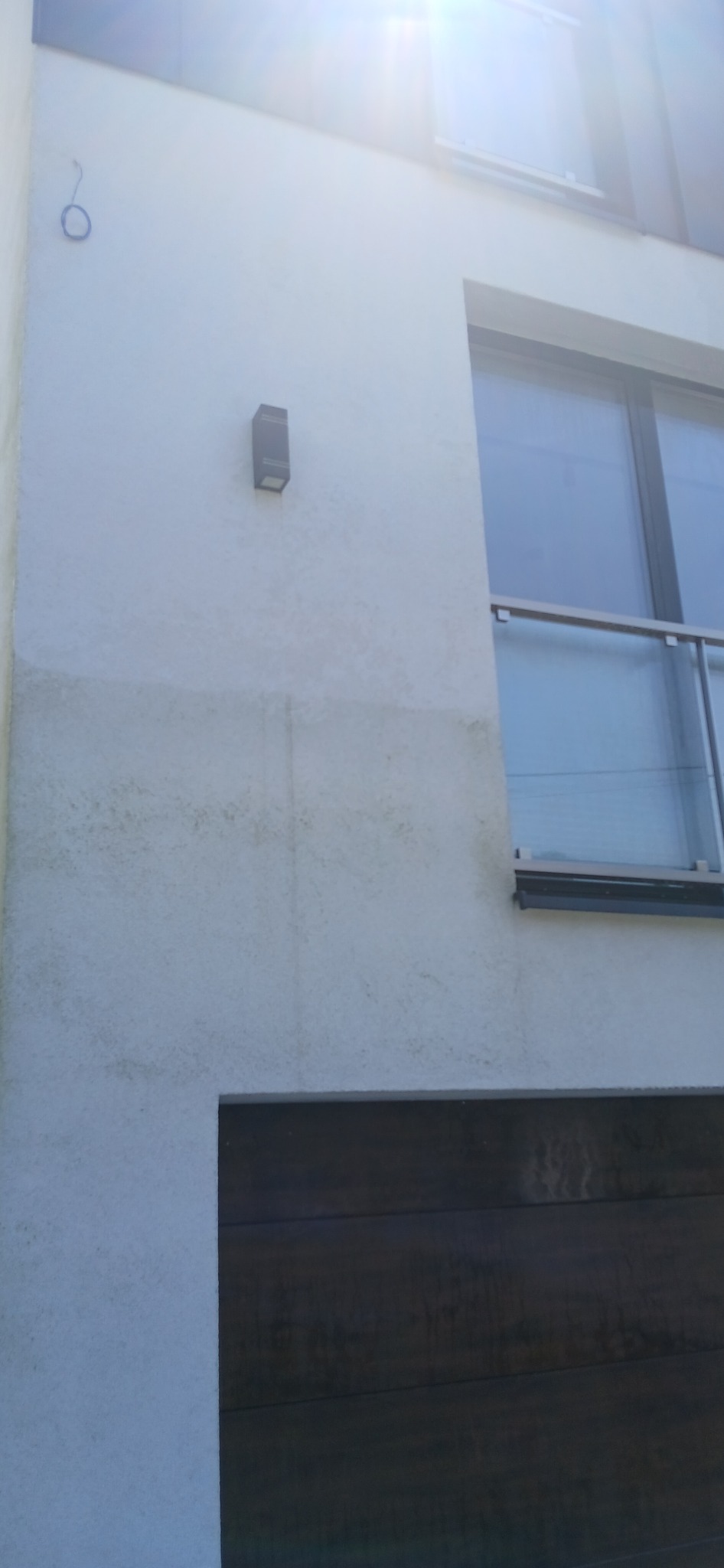 Boczna ściana budynku z widocznymi śladami zabrudzeń i zacieków na elewacji, fragment okna z barierką i brama garażowa z ciemnego drewna.