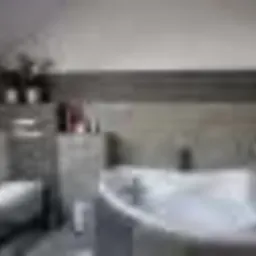 Jasna łazienka na poddaszu z białą wanną narożną, szarą glazurą i skośnym oknem dachowym. Widoczne akcesoria łazienkowe i toaleta. Całość utrzymana w nowoczesnym stylu.