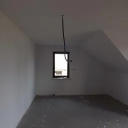 Poddasze po remoncie: świeżo pomalowane ściany na biało, okno w ramie z ciemnego drewna, widoczne przewody na suficie. Skośny sufit.