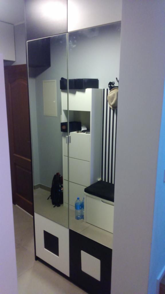 Szafa z lustrzanymi drzwiami w przedpokoju, odbijająca biały regał z siedziskiem, czarny plecak i butelki wody na podłodze.