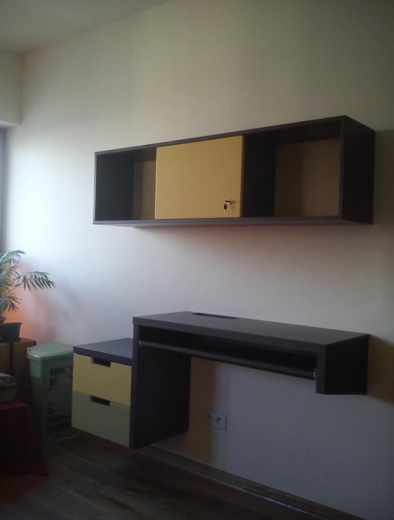 Wiszący zestaw biurkowy z szafką i szufladami w kolorach szaro-żółtych, zamek przy szafce, widoczne gniazdka elektryczne na ścianie, oświetlenie dzienne z lewej strony.