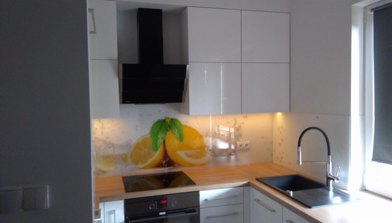 Nowoczesna, jasna kuchnia z białymi szafkami i drewnianym blatem, ozdobiona panelem szklanym z motywem cytryn, czarny okap nad płytą indukcyjną, zlewozmywak z wysoką baterią przy oknie.