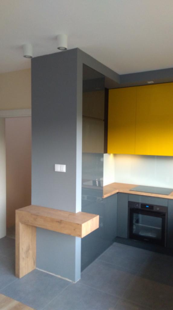 Nowoczesna kuchnia z szarymi szafkami dolnymi, drewnianym blatem, żółtymi szafkami górnymi i dekoracyjnym półwyspem z drewnianym blatem.