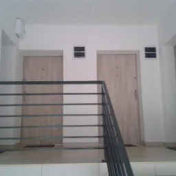 Klatka schodowa z dwoma jasnymi, drewnopodobnymi drzwiami, widoczne zza metalowej balustrady, z elementami instalacji na ścianach.