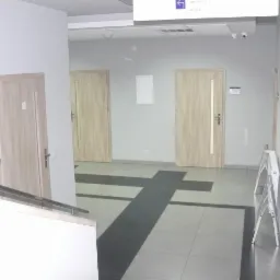 Korytarz w budynku użyteczności publicznej z trzema parami drzwi drewnianych o jasnej kolorystyce i szarą podłogą z wzorem w kształcie krzyża.