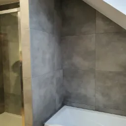 Nowoczesna łazienka w Przelewicach po remoncie: szare, matowe płytki wielkoformatowe na ścianach, biała wanna akrylowa i kabina prysznicowa ze szklanymi drzwiami.