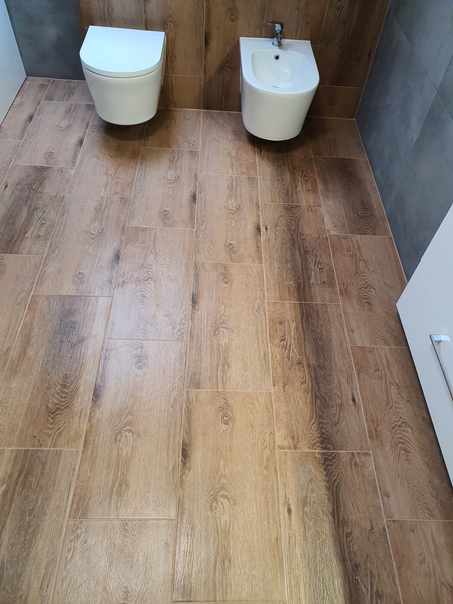 Nowoczesna łazienka z drewnopodobną podłogą, wiszącą toaletą i bidetem, szare płytki na ścianie w tle.