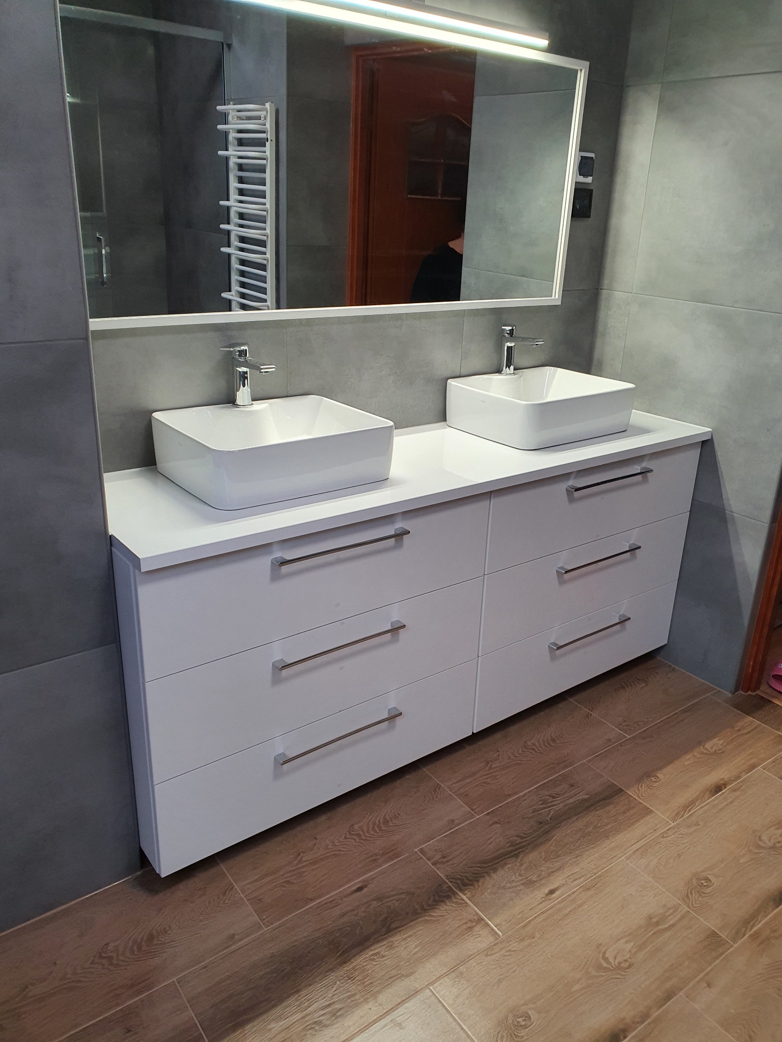 Nowoczesna łazienka z dwiema kwadratowymi umywalkami nablatowymi na białej szafce z sześcioma szufladami, szare płytki na ścianach, drewnopodobna podłoga i lustro z oświetleniem LED.