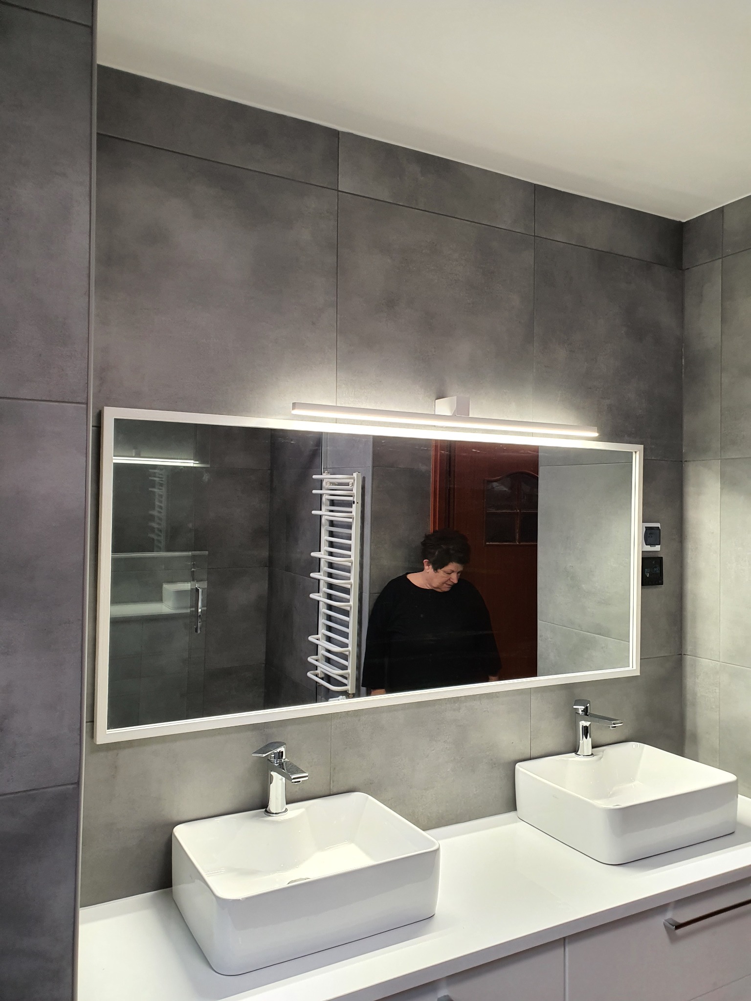 Nowoczesna łazienka z dwoma białymi umywalkami nablatowymi, dużym lustrem z oświetleniem LED i szarymi płytkami na ścianach, widoczne odbicie osoby.