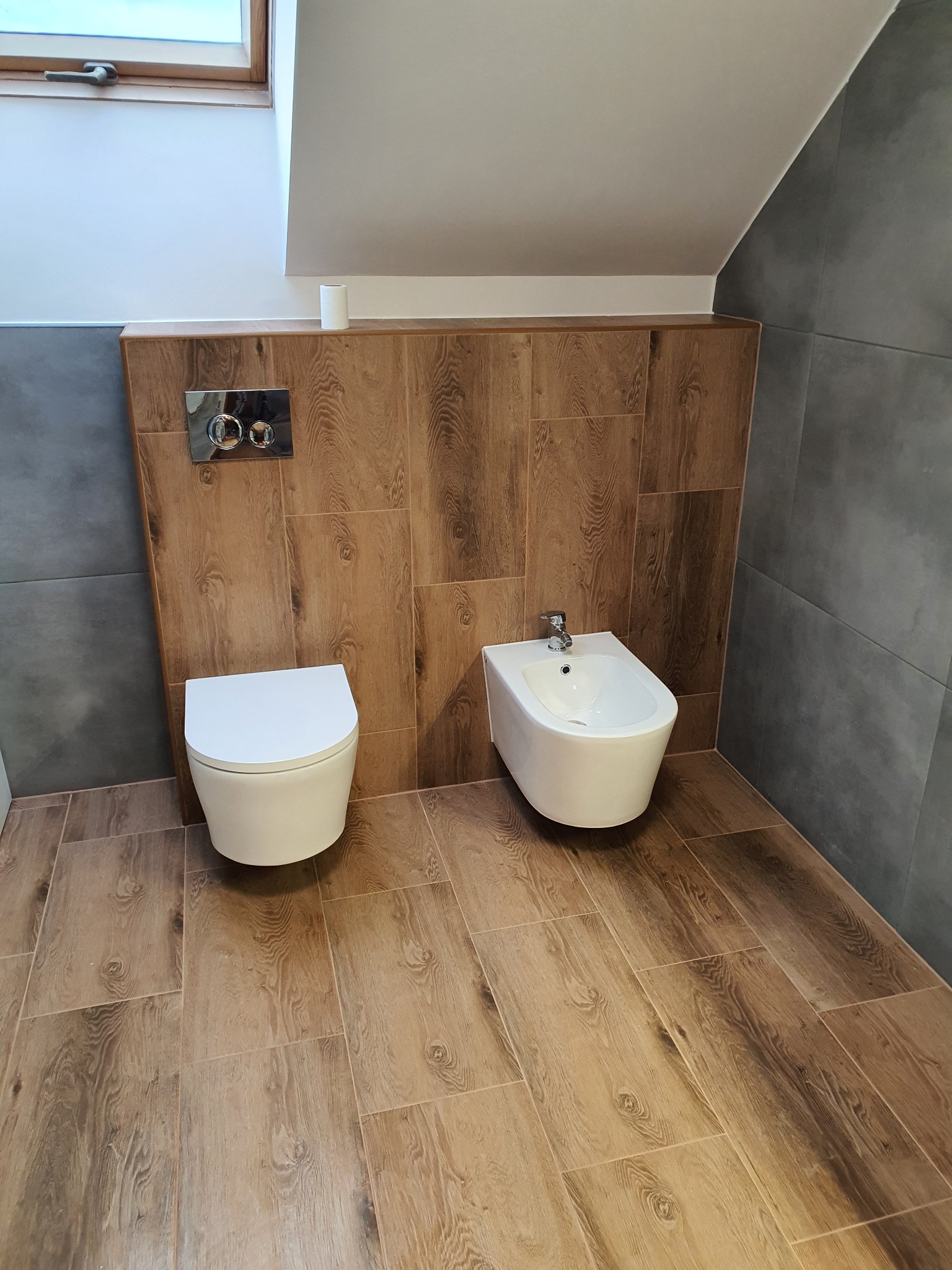 Nowoczesna łazienka na poddaszu z drewnopodobną podłogą i ścianą, wisząca toaleta i bidet z chromowaną armaturą, szare płytki na ścianie bocznej, okno dachowe.