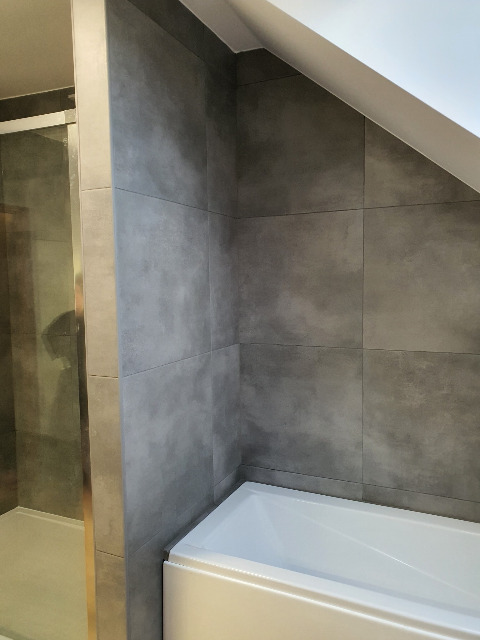 Nowoczesna łazienka w Przelewicach po remoncie: szare, matowe płytki wielkoformatowe na ścianach, biała wanna akrylowa i kabina prysznicowa ze szklanymi drzwiami.